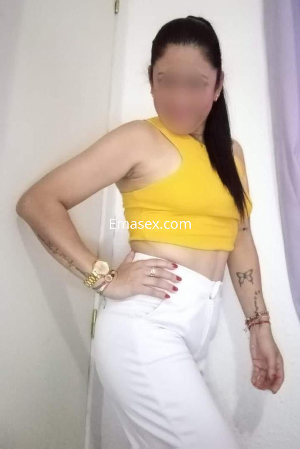 Escort en Castellón con blusa amarilla sin mangas y pantalones blancos, posando con confianza y mostrando sus tatuajes.