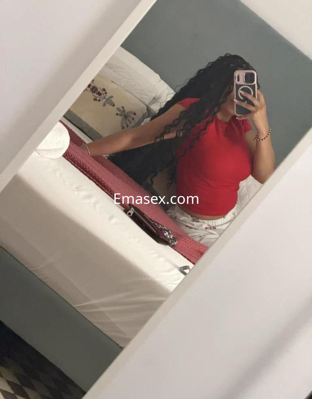 Puta en Granada, con cabello rizado y largo, viste una camiseta roja y pantalones cortos, posando frente al espejo.