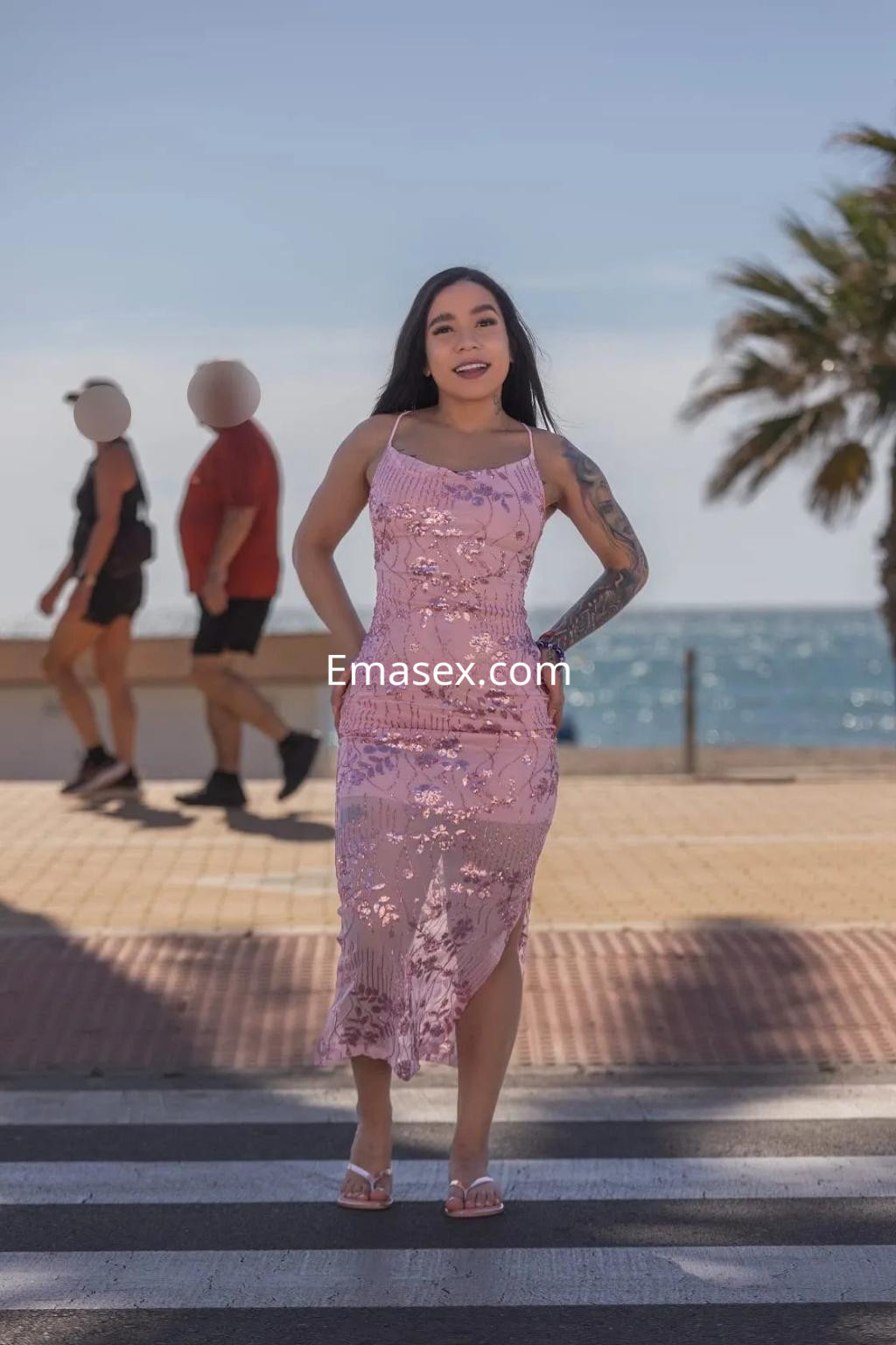 Puta posando con un vestido rosa con detalles florales en una playa de Granada, con palmeras al fondo.