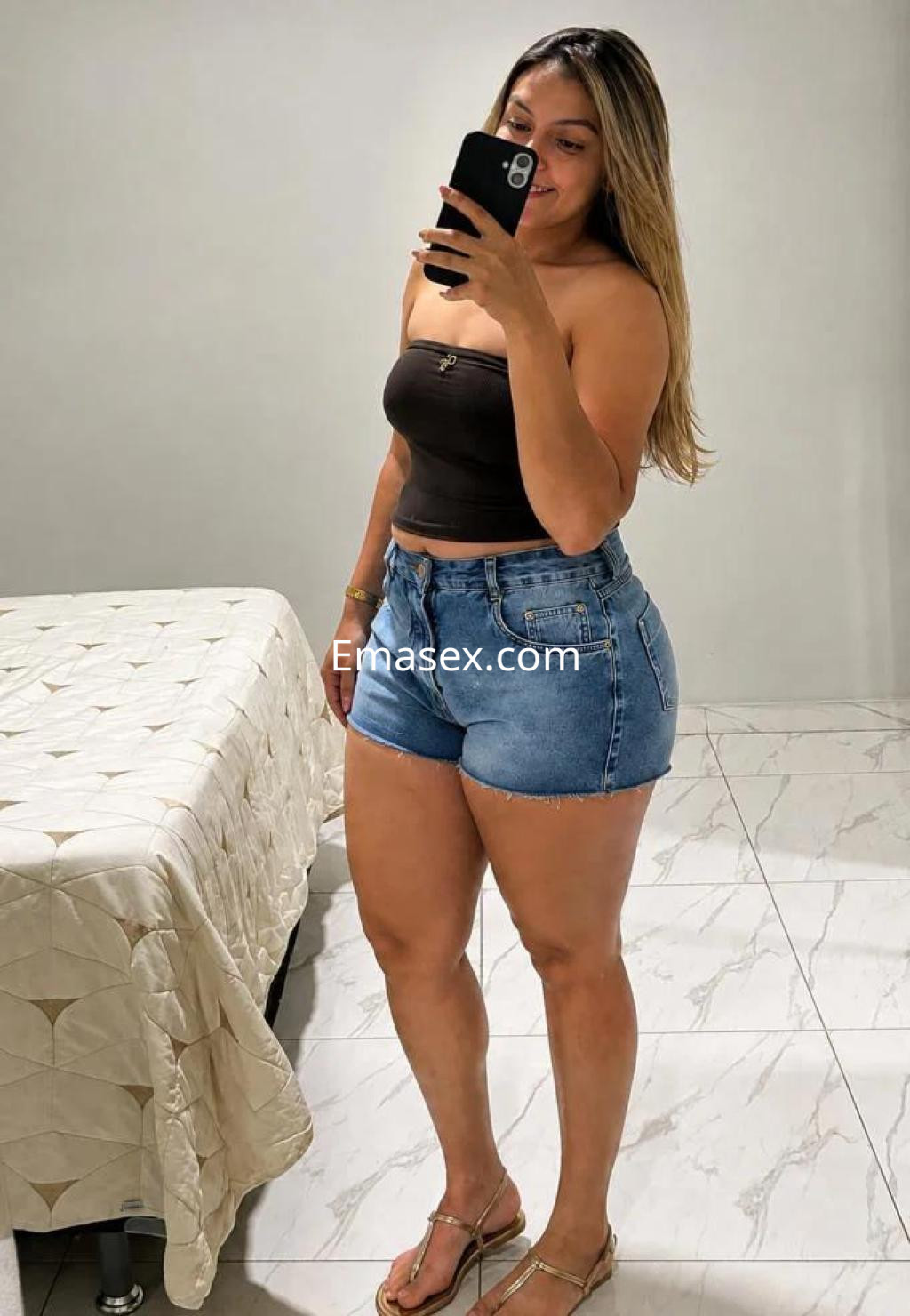 Puta en Huelva se muestra frente al espejo, vistiendo un top oscuro y shorts de mezclilla, con cabello largo y lacio.