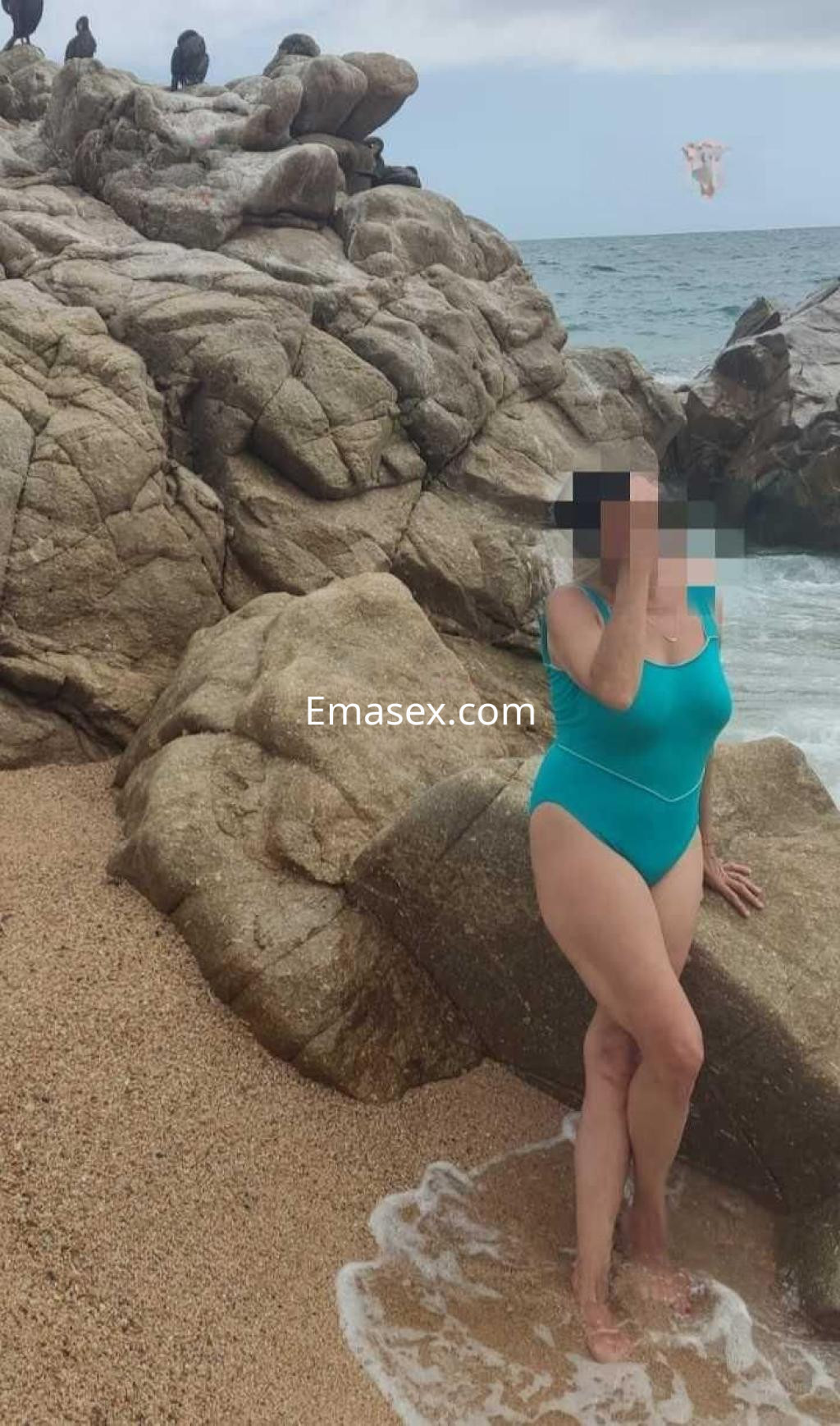 Puta de Lugo posando en la playa, vestida con un traje de baño azul, junto a rocas y el mar de fondo.