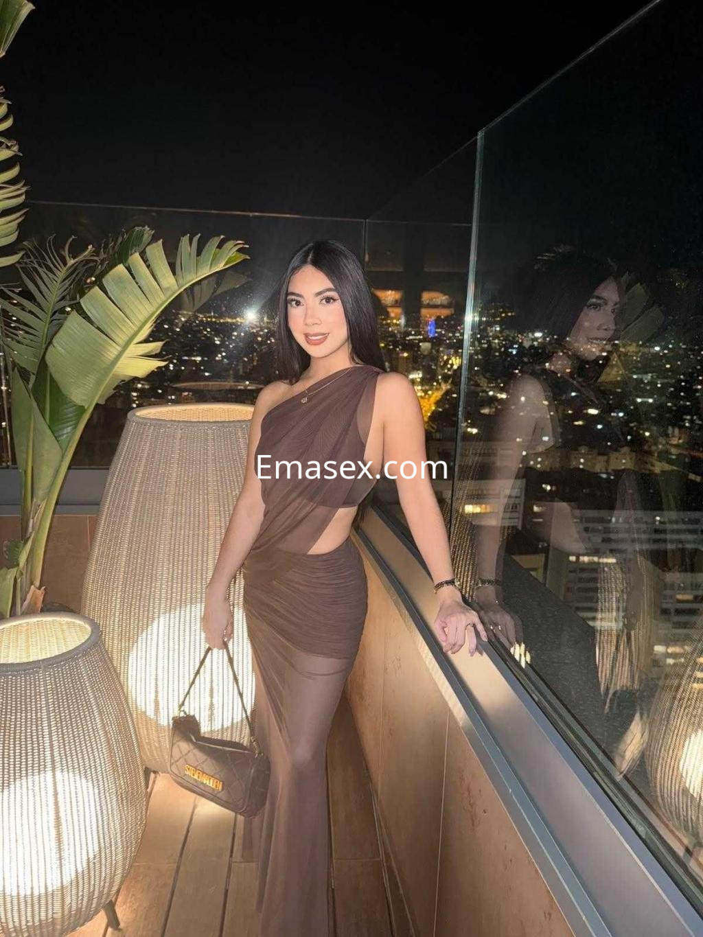 Puta en Madrid posando en una terraza iluminada, vestida con un vestido largo y transparente de color marrón.