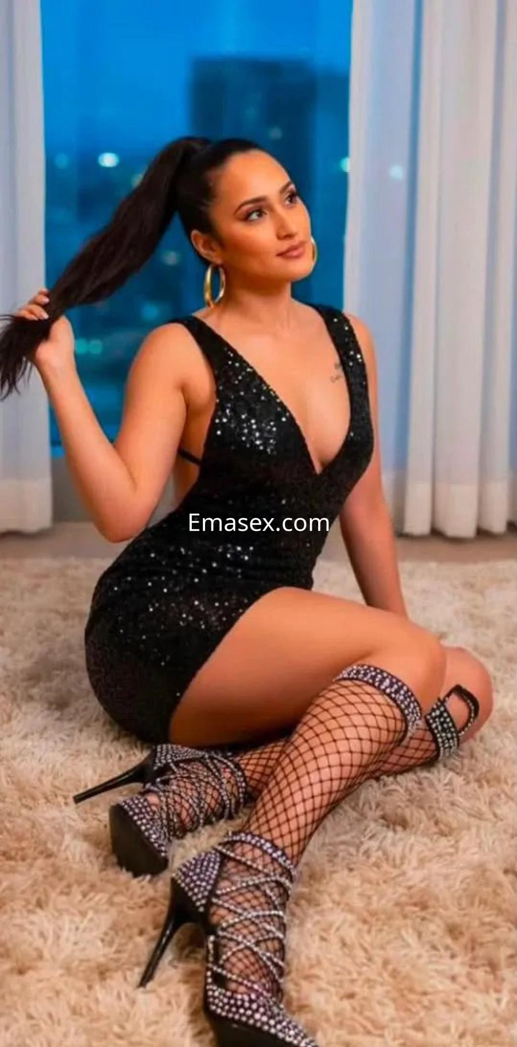 Escort en Madrid luciendo un vestido negro con brillos, mallas de red y tacones, posando sentada en un ambiente elegante.