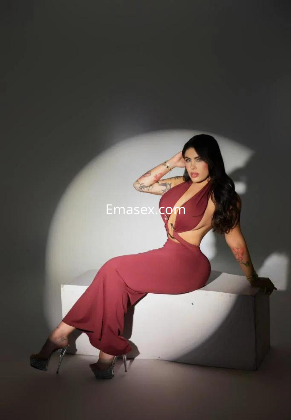 Escort en Sevilla posa con un vestido largo de color burdeos, sentada sobre un soporte blanco, luciendo un peinado ondulado.