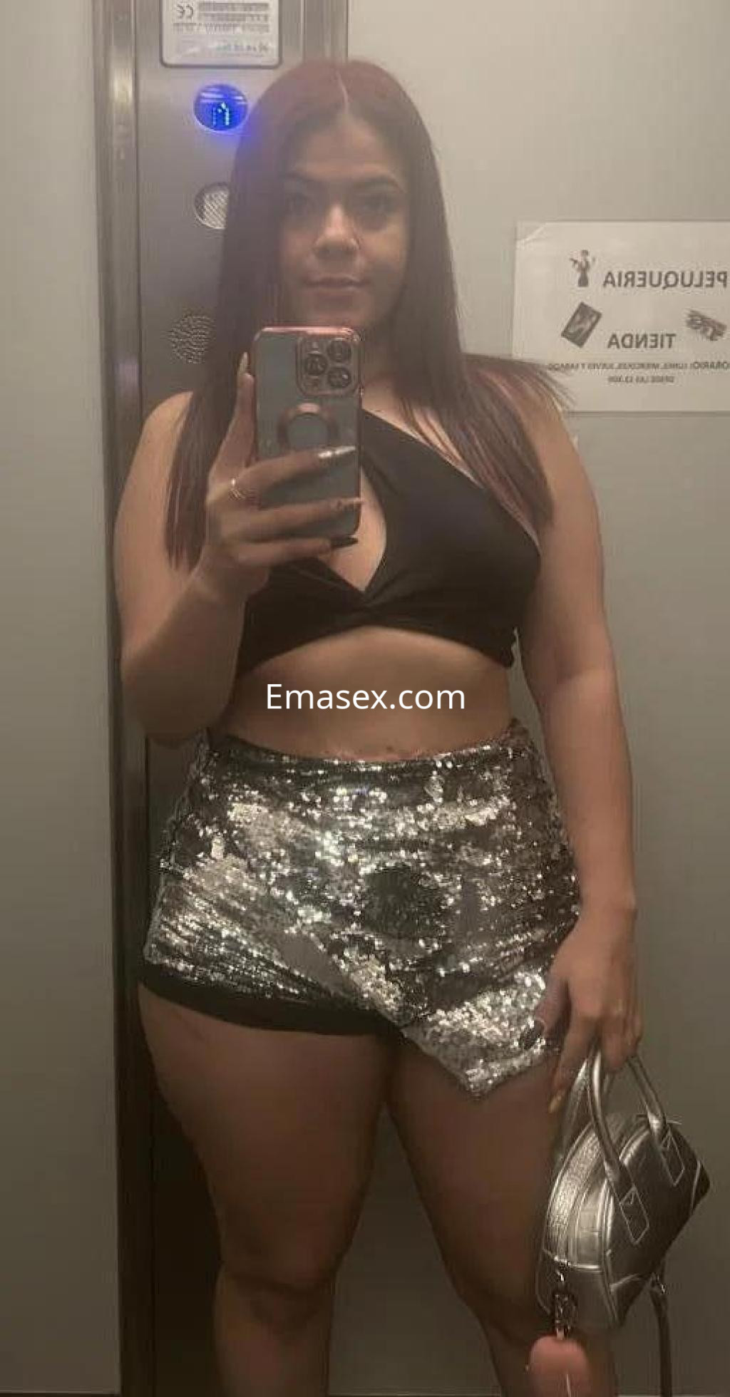 La puta en Sevilla se muestra en un ascensor, luciendo una blusa negra y shorts brillantes con un bolso plateado.