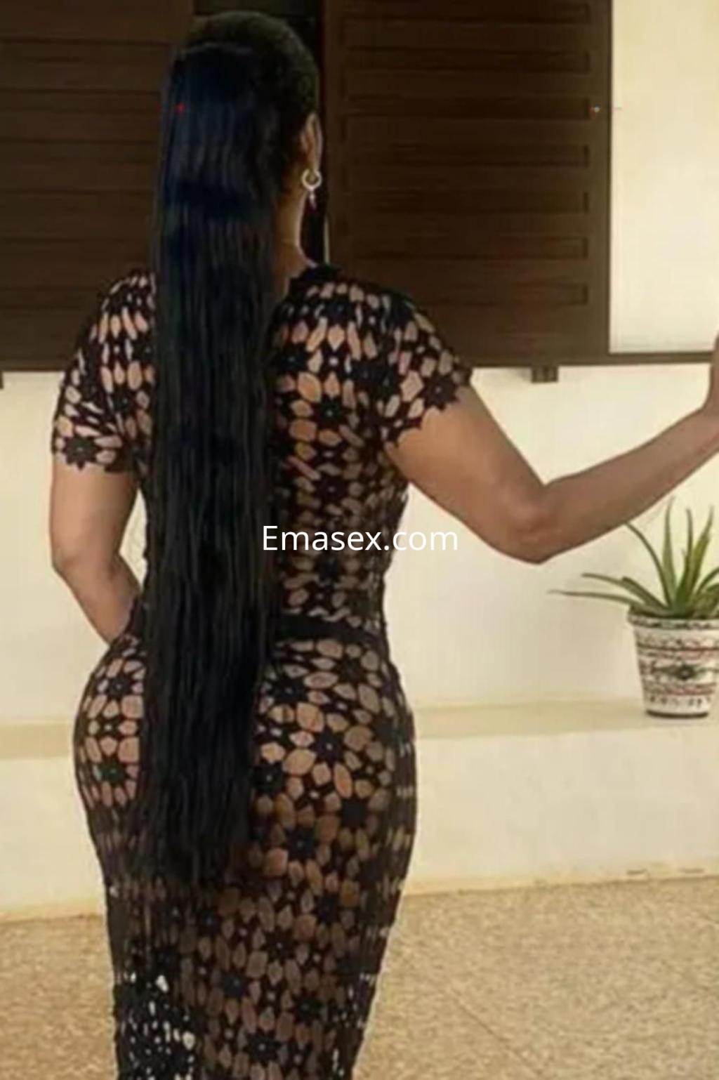 Escort en Sevilla con vestido de encaje negro, mostrando su largo cabello y posando de espaldas en un entorno exterior.