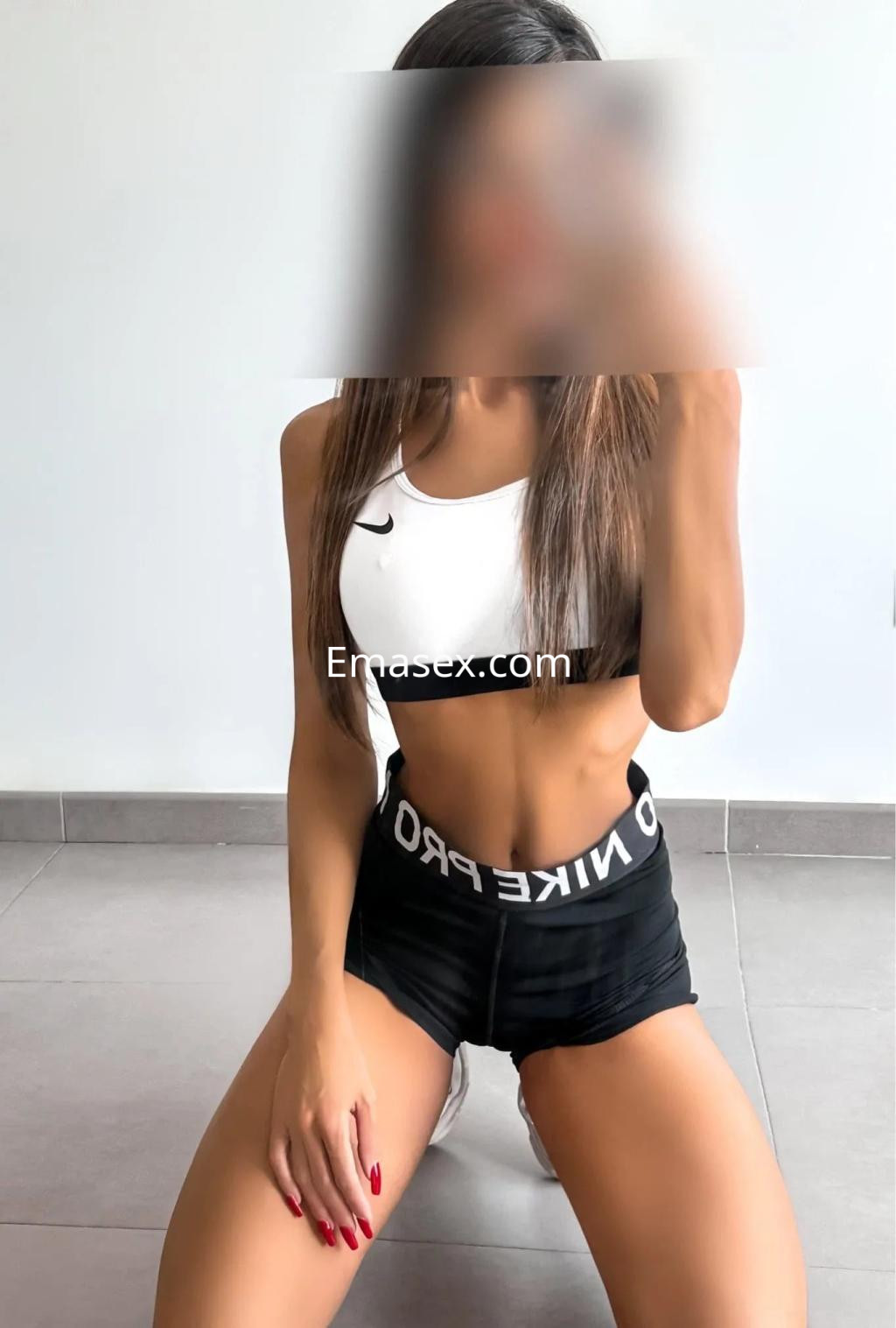 Escort en Valencia con top blanco y pantalones cortos negros, posando de rodillas en un entorno minimalista.