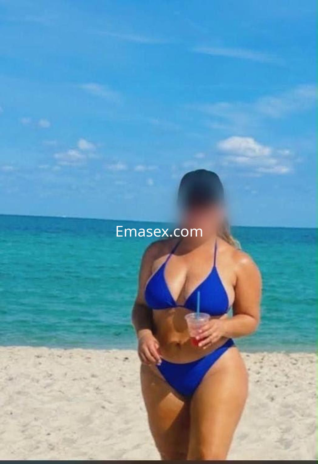 Escort en Vitoria, caminando por la playa con un bikini azul y sosteniendo una bebida refrescante.