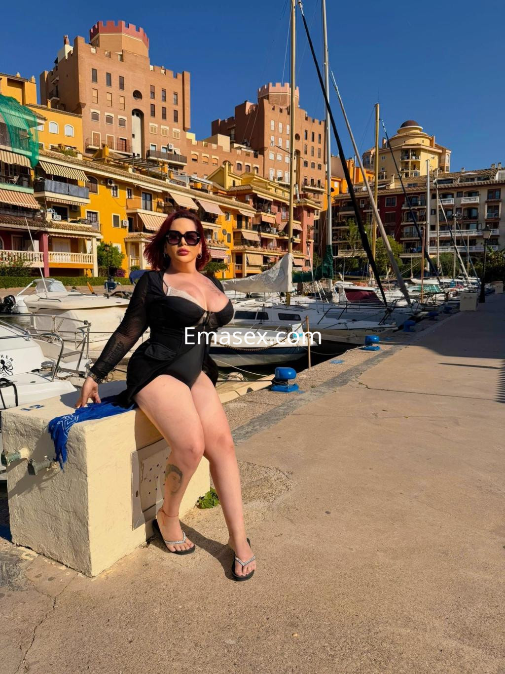 Escort en Alicante vistiendo un traje de baño negro y sandalias, posando con confianza junto al puerto.