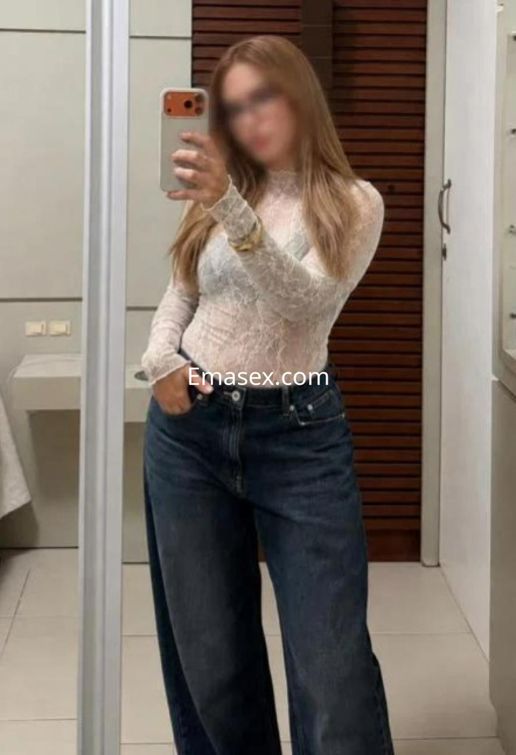 Escort en Barcelona con cabello largo y rubio, posando en un espejo con blusa de encaje y jeans amplios.