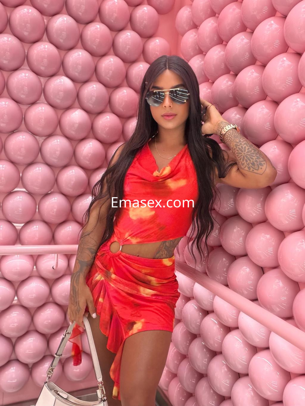 Escort en Tenerife con cabello largo y lacio, viste un conjunto rojo con estampado y accesorios elegantes, posando entre paredes de color rosa.