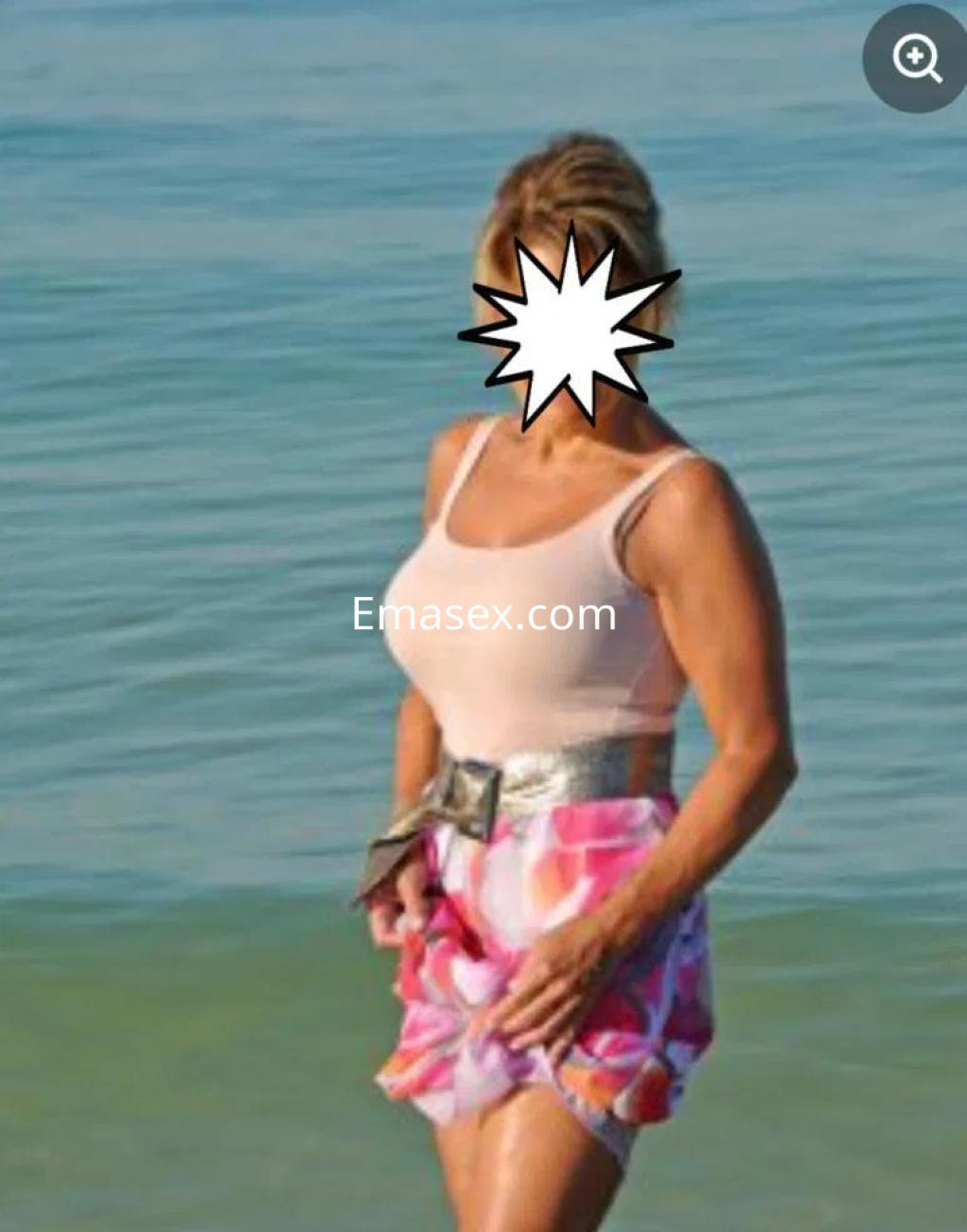 Escort en Alicante, con cabello rubio y piel bronceada, luciendo un vestido de baño claro y una falda colorida en la playa.