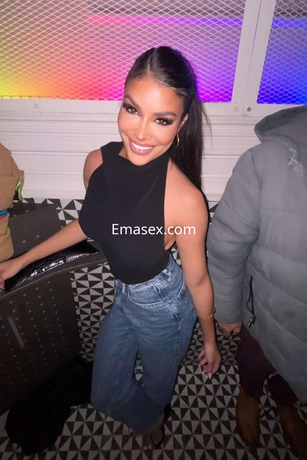 Escort en Ibiza con cabello largo y oscuro, vistiendo blusa sin mangas y jeans, sonriendo en un ambiente festivo.