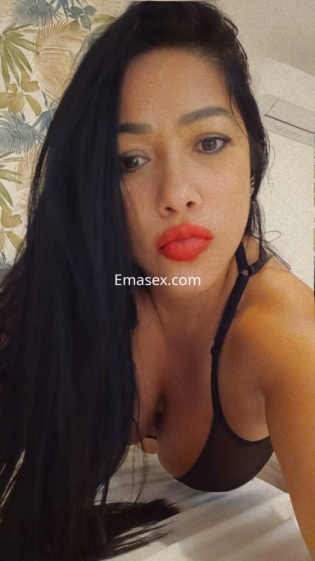 La puta posa en lencería negra, con labios rojos y cabello largo, en un entorno con fondo floral en Barcelona.