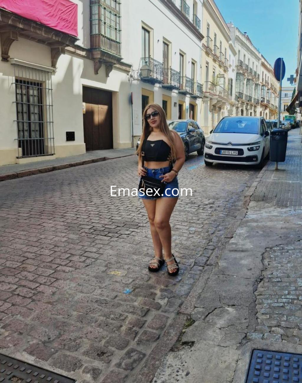 Puta posando en una calle de Cáceres, lleva un top negro, shorts de mezclilla y gafas de sol.