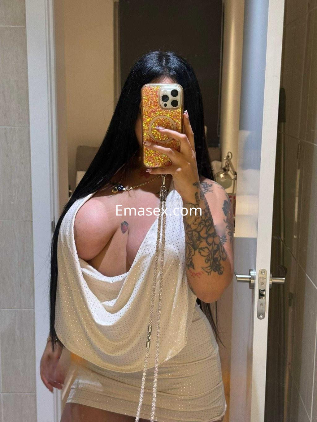 Escort en Huelva posando con un vestido blanco brillante, mostrando su cabello largo y oscuro y tatuajes en los brazos.