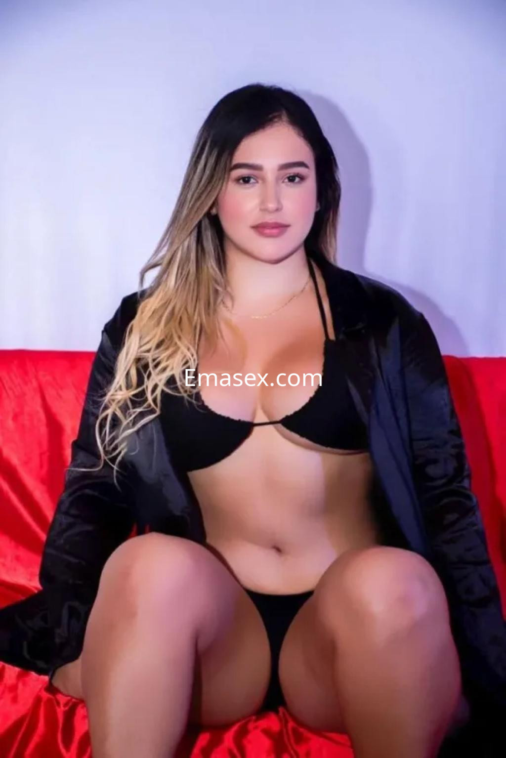 La puta posa sentada sobre un sofá rojo, vistiendo un bikini negro y una chaqueta oscura, con cabello largo y lacio.