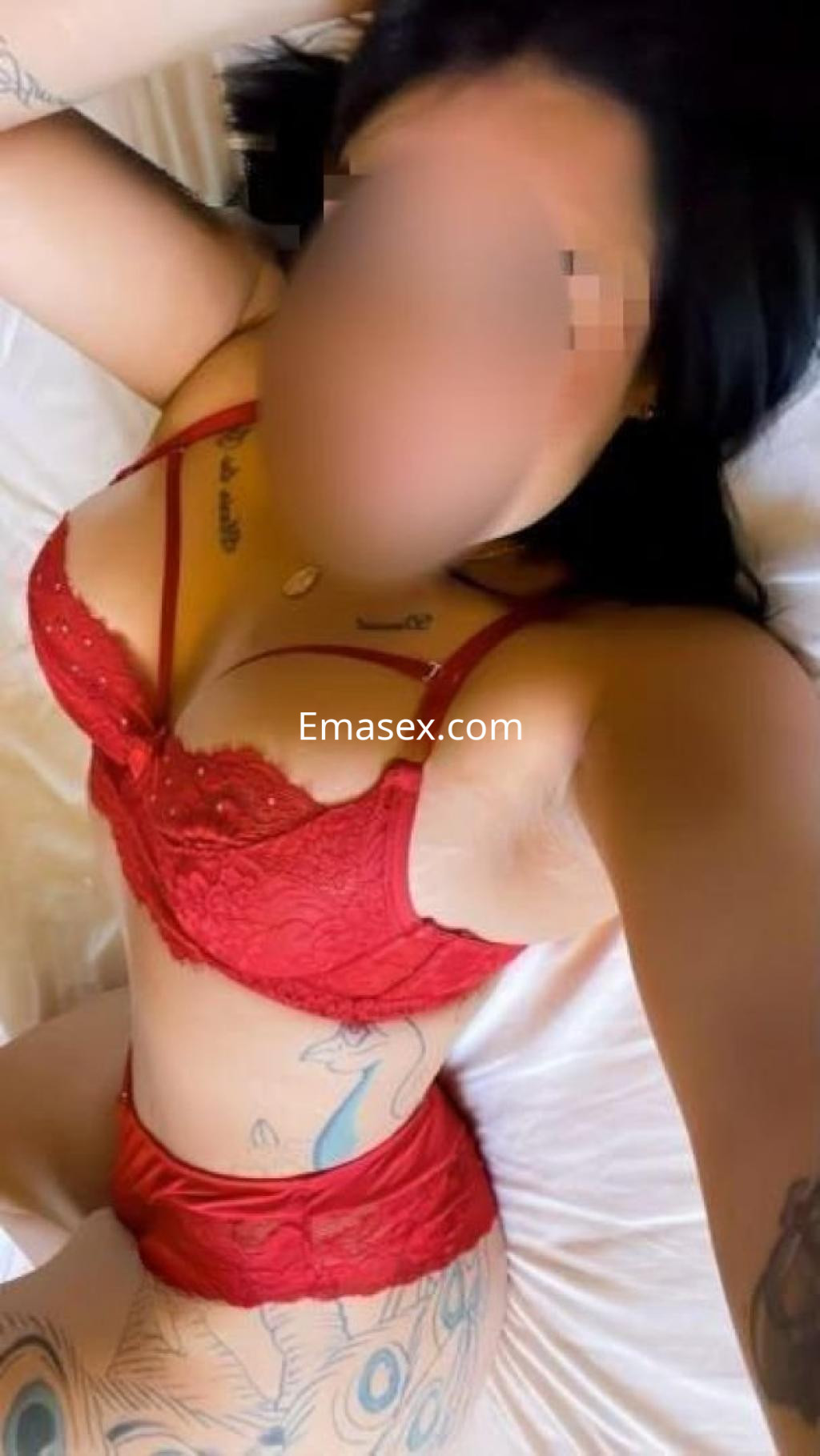 Escort en Lugo posando sobre la cama con lencería roja, mostrando sus tatuajes y un peinado de cabello oscuro.