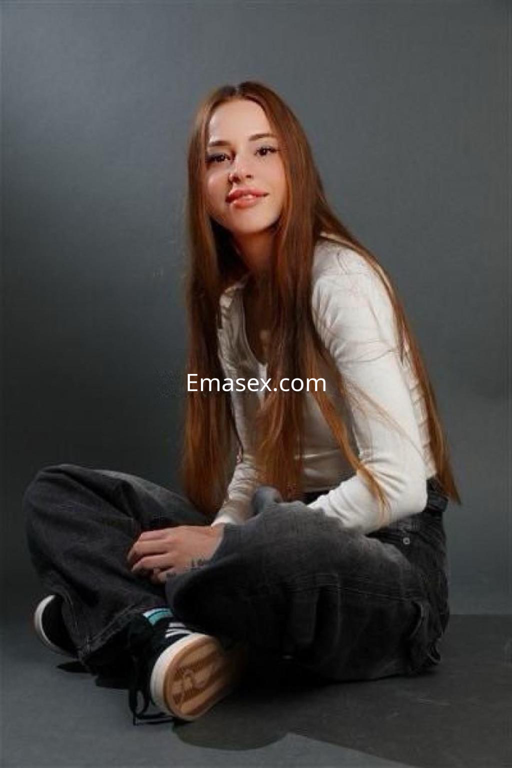 Escort en Madrid con cabello largo y lacio, vistiendo una camiseta blanca y pantalones oscuros, posando sentada.