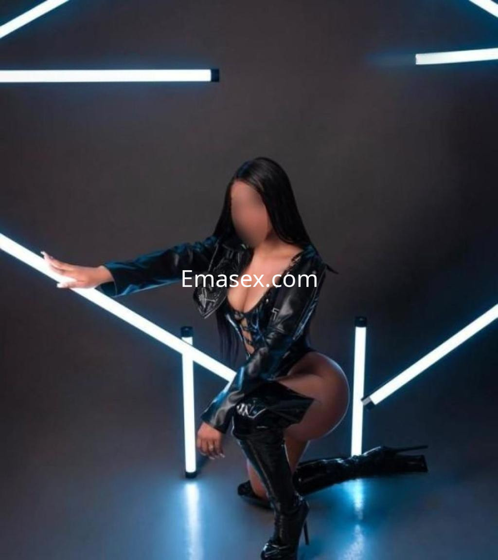 Puta de cabello largo y oscuro, vestida con un body negro y botas altas, posando en un fondo con luces en Madrid.