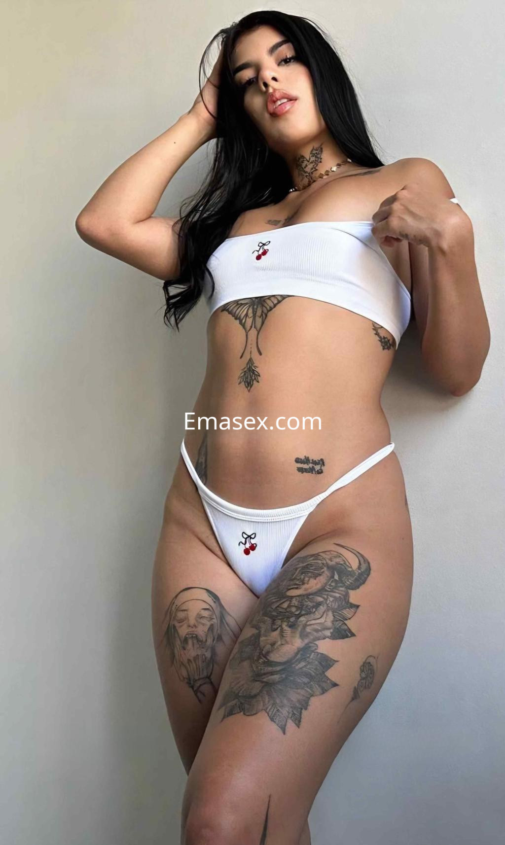 Escort en Madrid con piel clara, cabello largo y lacio, luciendo un top blanco y una tanga, exhibiendo varios tatuajes.