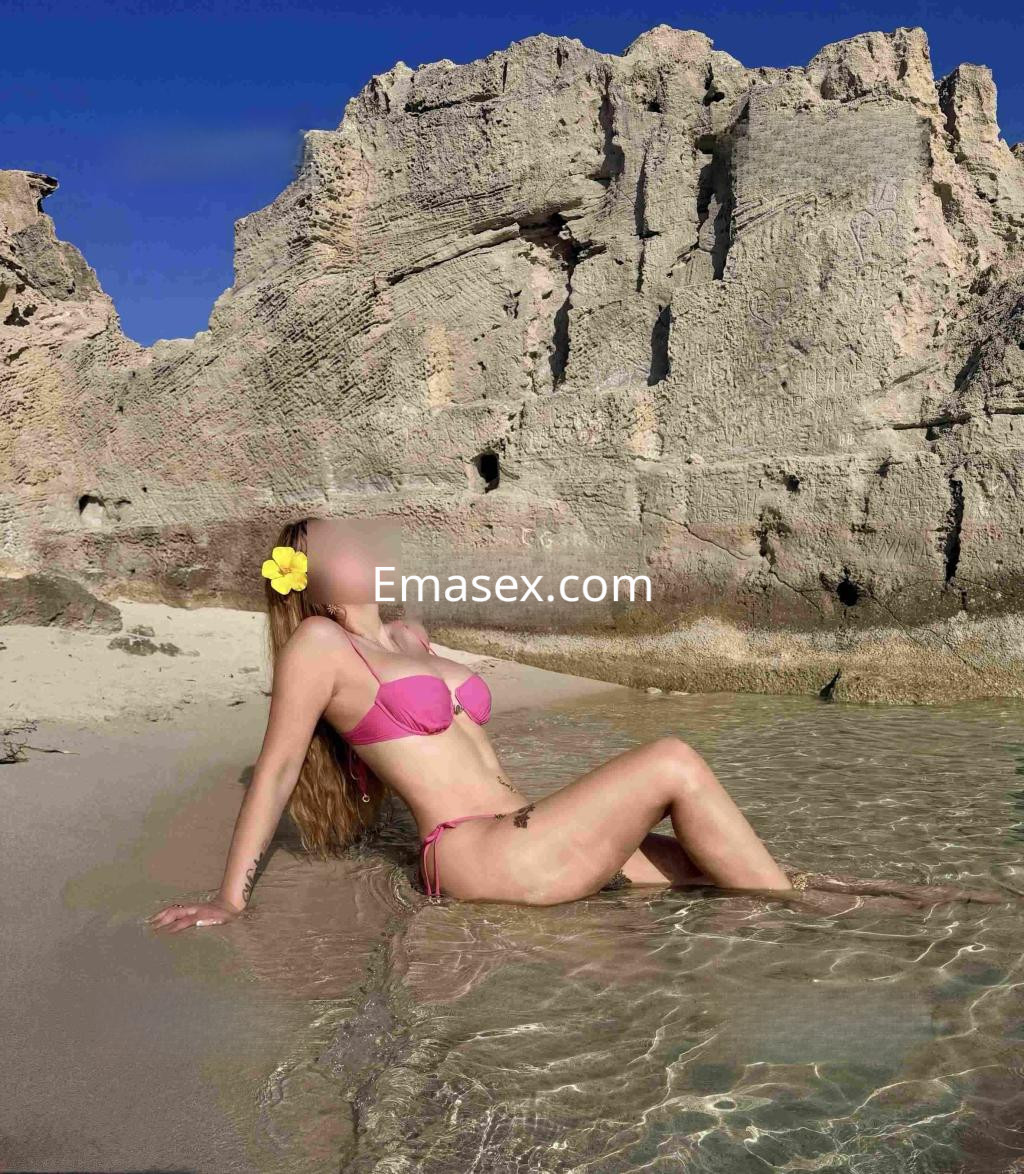 Escort en Madrid, con cabello largo y rubio, posa en la playa con bikini rosa, disfrutando del agua y el paisaje.