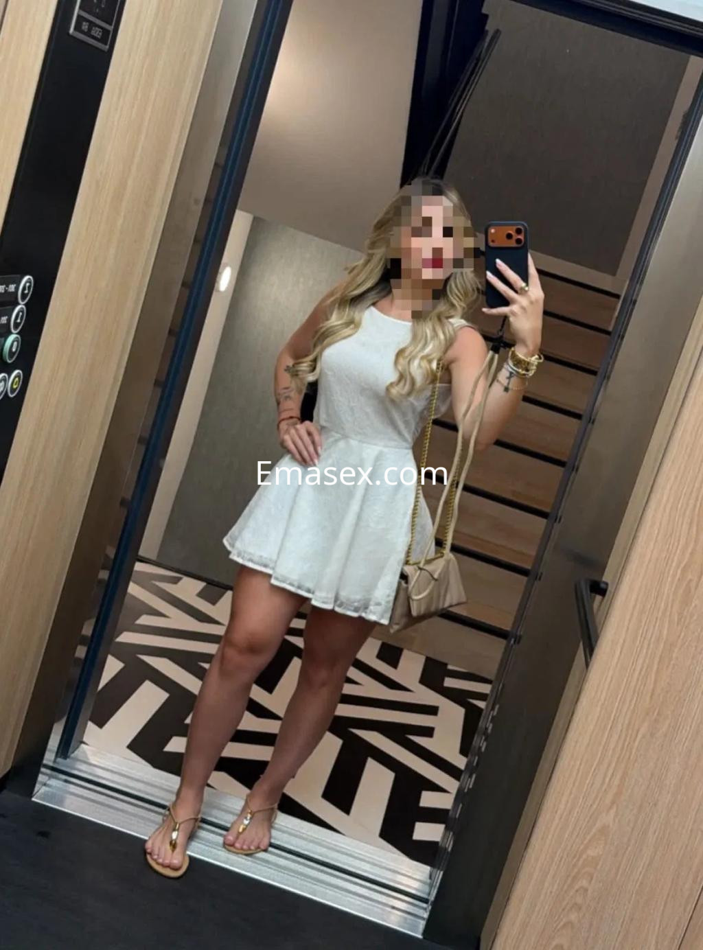 Puta en Murcia posa en un ascensor, vistiendo un vestido blanco y sandalias, con cabello largo y ondulado.