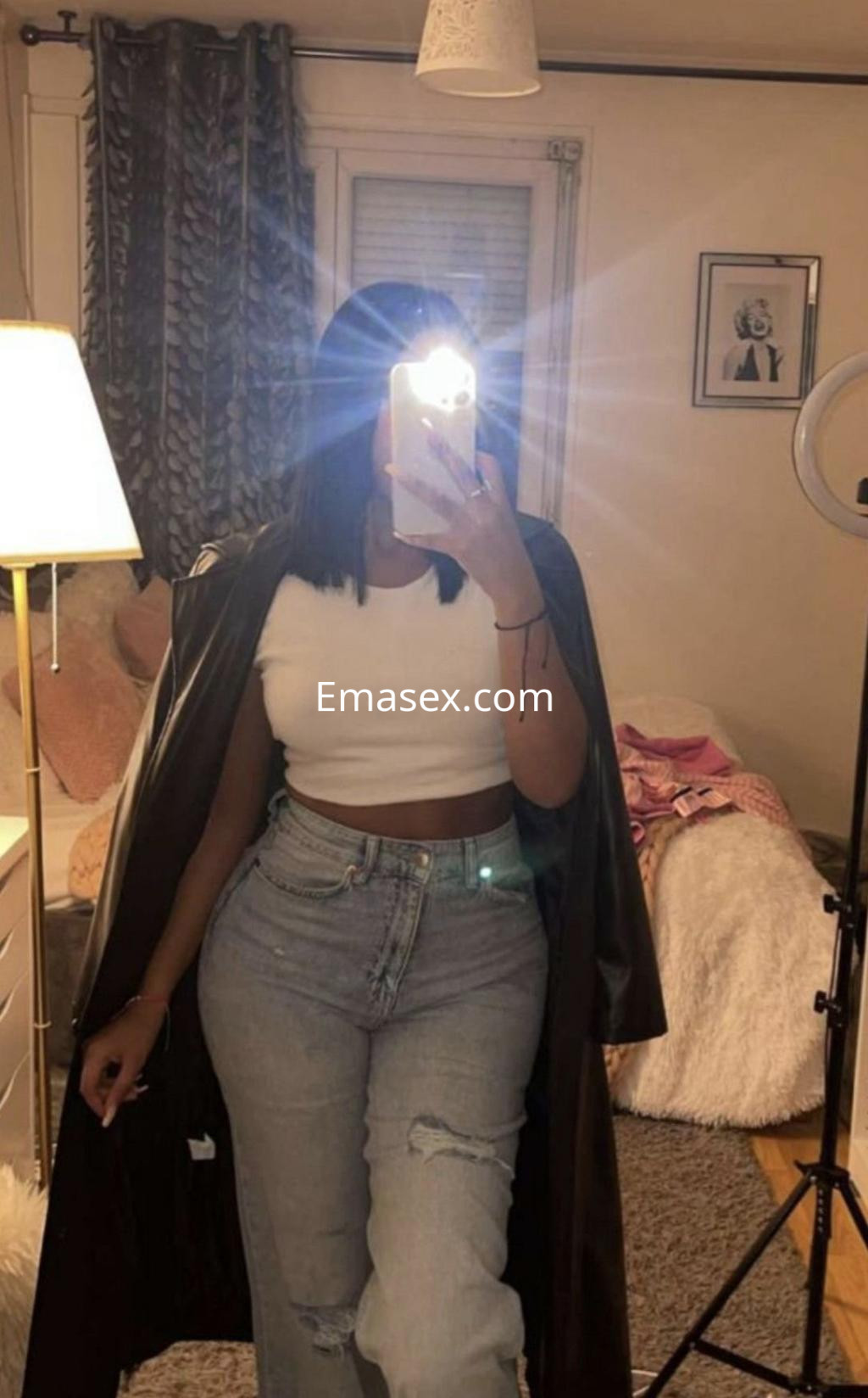 Escort en Murcia viste una blusa blanca y jeans ajustados, posando con una chaqueta negra en un ambiente acogedor.