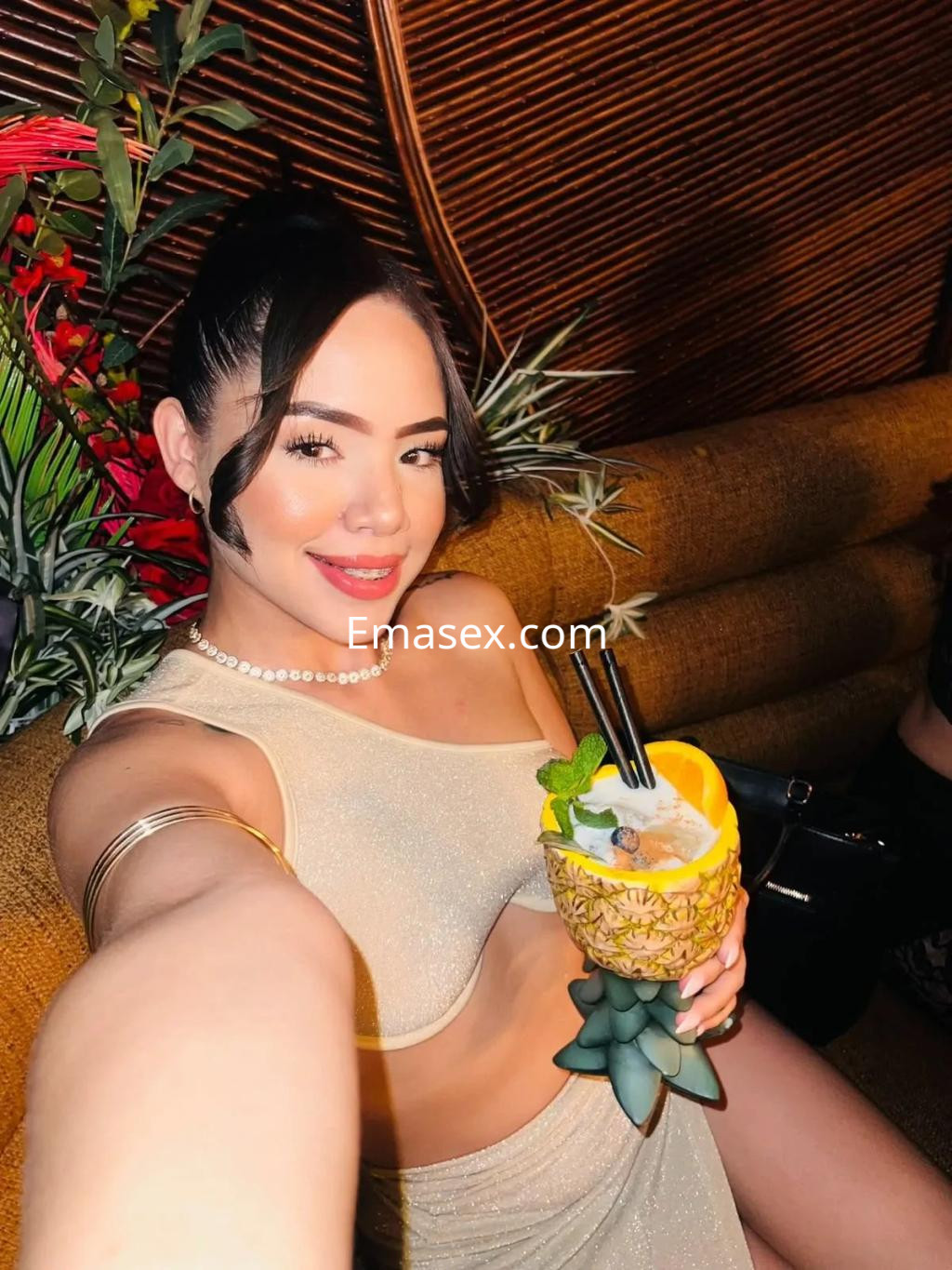 Puta sonriente en Vigo, con top dorado y cóctel servido en piña, posando en un ambiente tropical.
