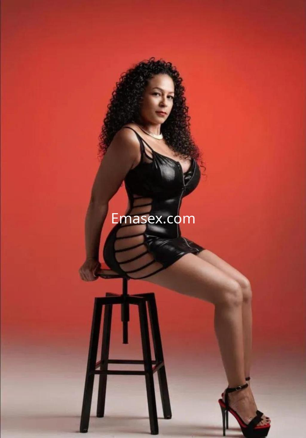 Escort en Tenerife con cabello rizado y vestido de cuero oscuro, posando sentada sobre un taburete.