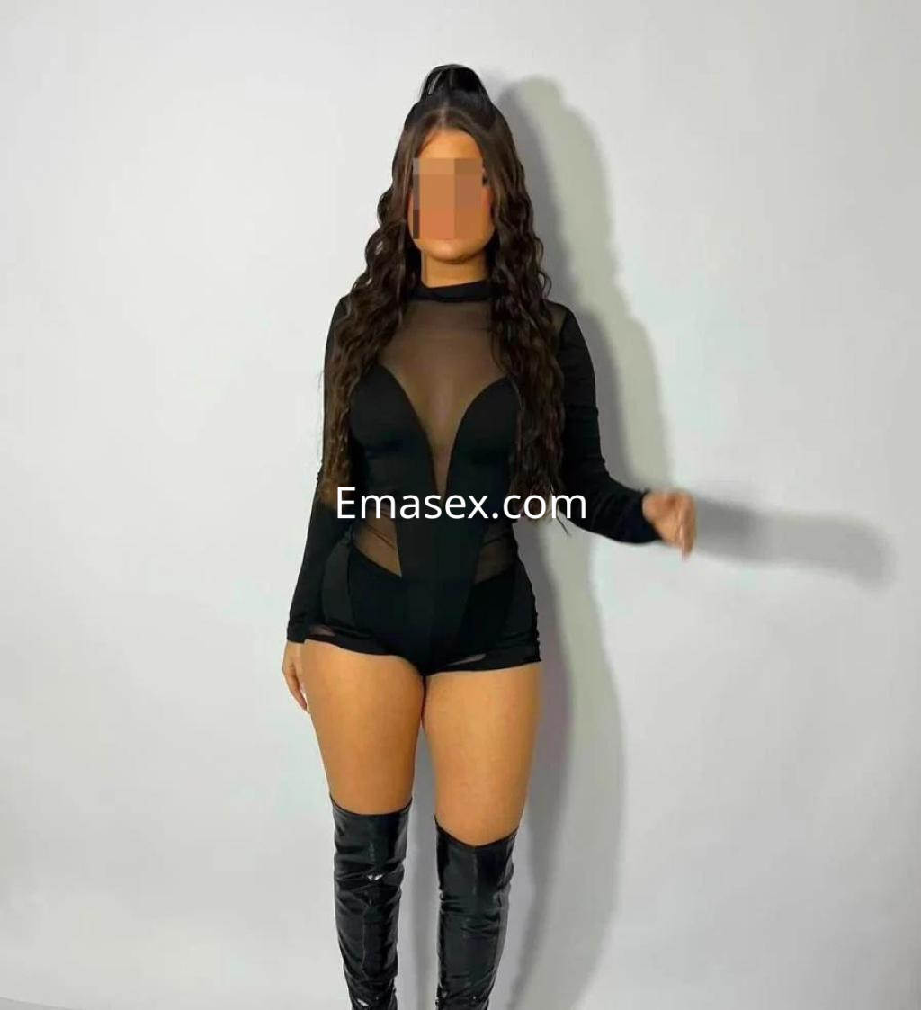 Escort en Tenerife luciendo un atuendo negro ajustado con transparencias y botas largas, posando frente a un fondo neutro.
