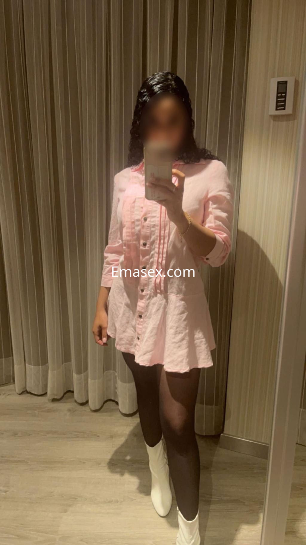 Escort en Valencia viste una blusa rosa con botones, falda corta y botas blancas, posando frente al espejo.