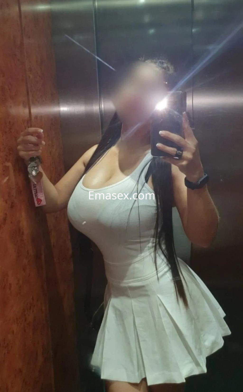 Escort en Málaga, posando en un ascensor con vestido blanco ajustado y cabello largo, sosteniendo su teléfono.