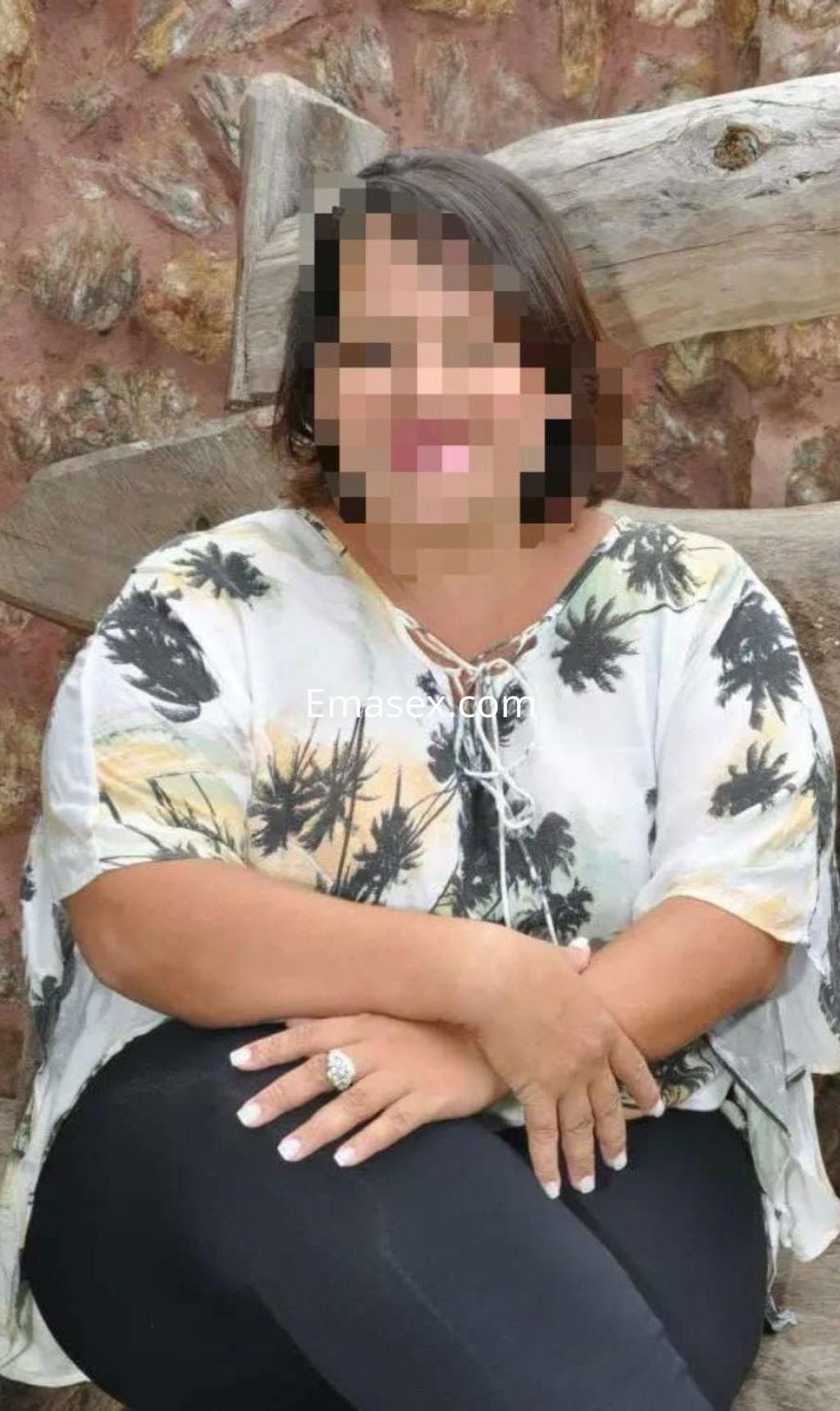 Puta sentada en un banco de madera, vistiendo blusa ligera con motivos tropicales y pantalones oscuros en Oviedo.
