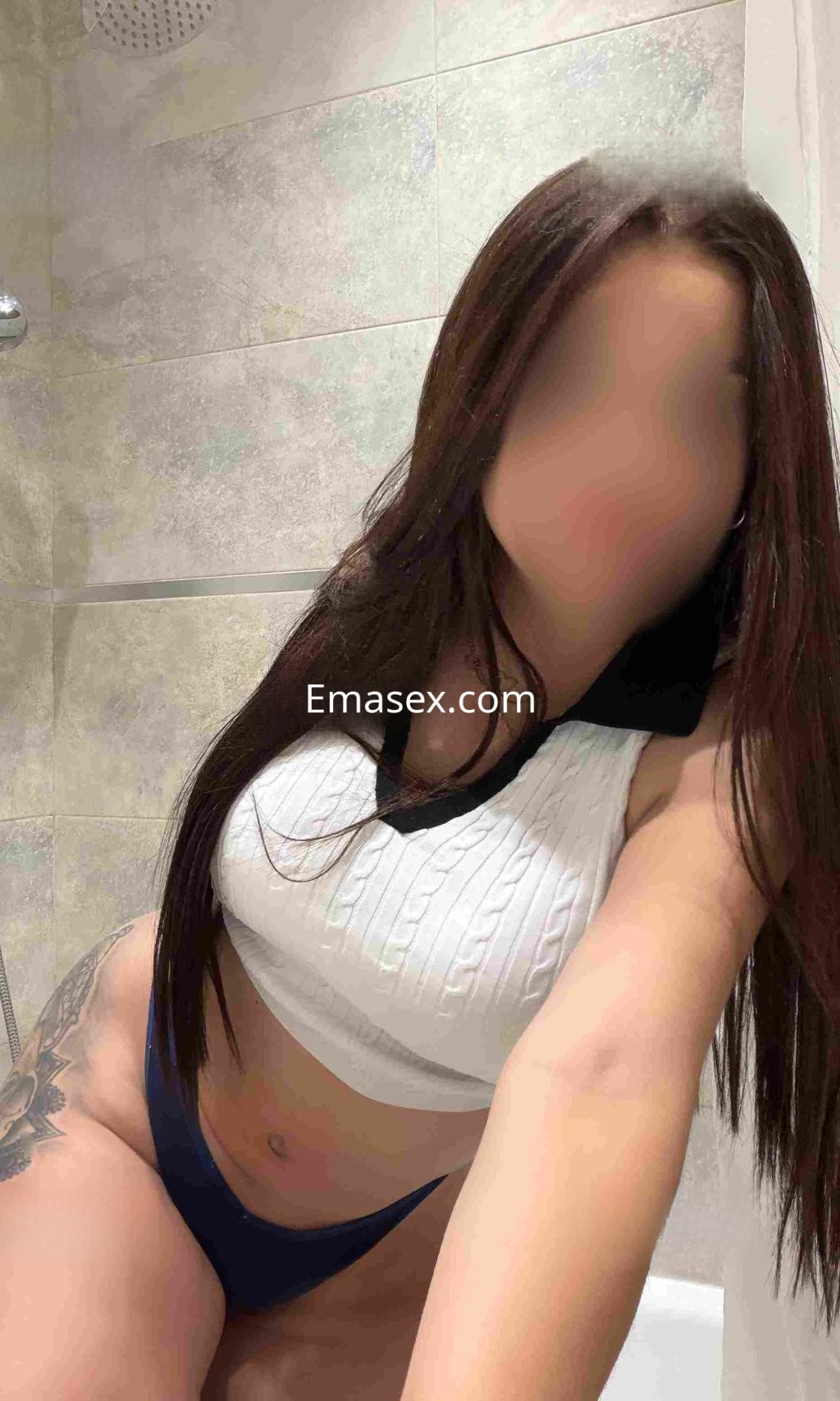Escort en Valladolid, con cabello largo y lacio, viste un top blanco y braguitas oscuras, posando en un baño.