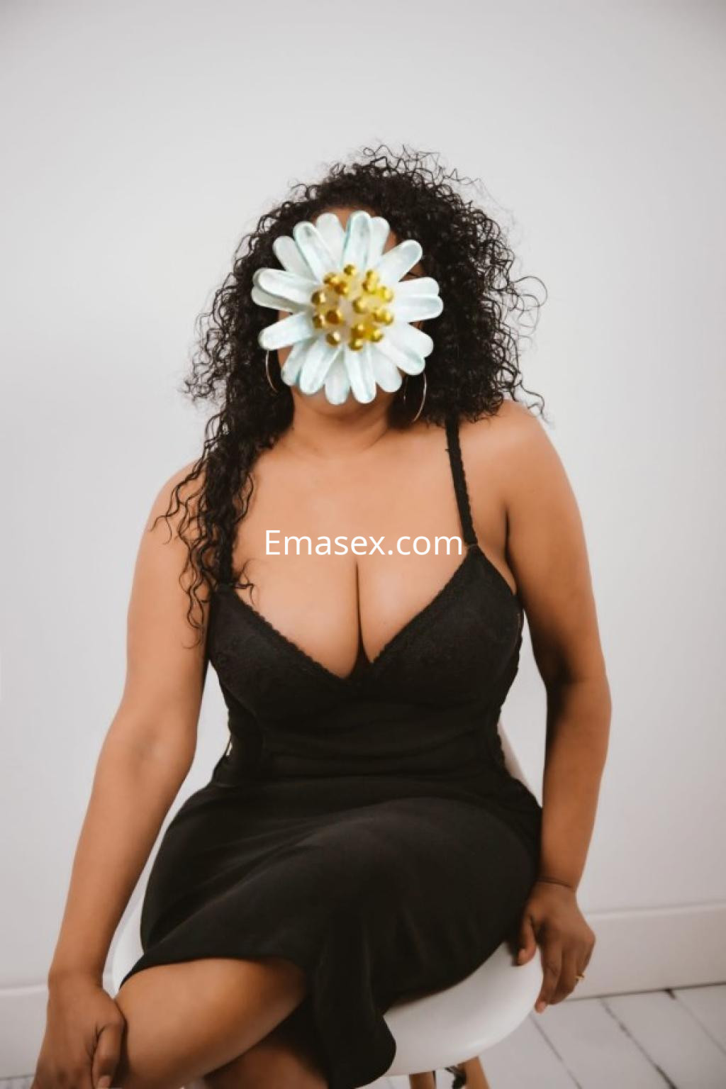 Escort en Oviedo posando sentada con un vestido negro, cabello rizado y una flor cubriendo su rostro.