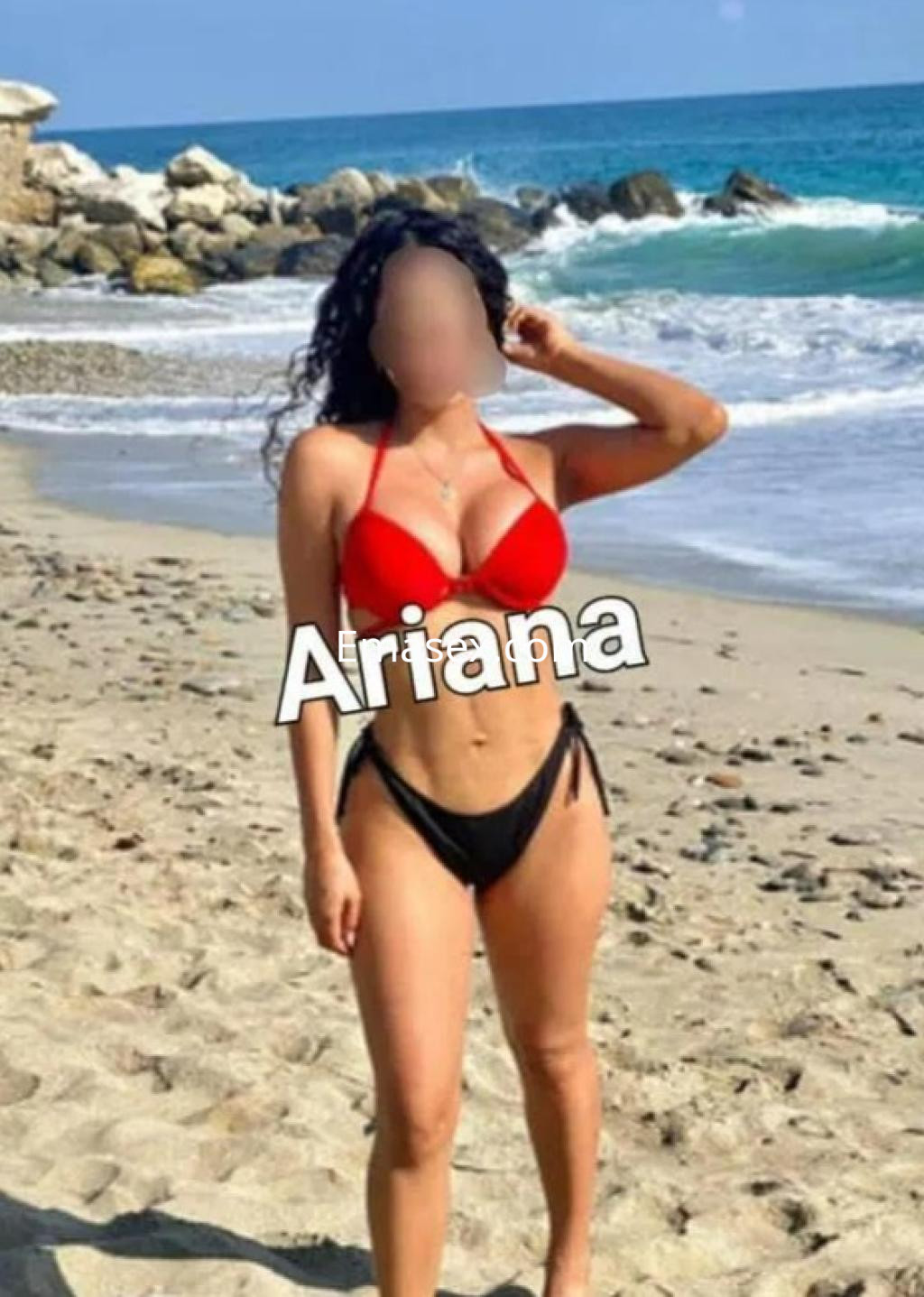 Escort en Barcelona luciendo un bikini rojo y negro en la playa, con el mar y rocas al fondo.