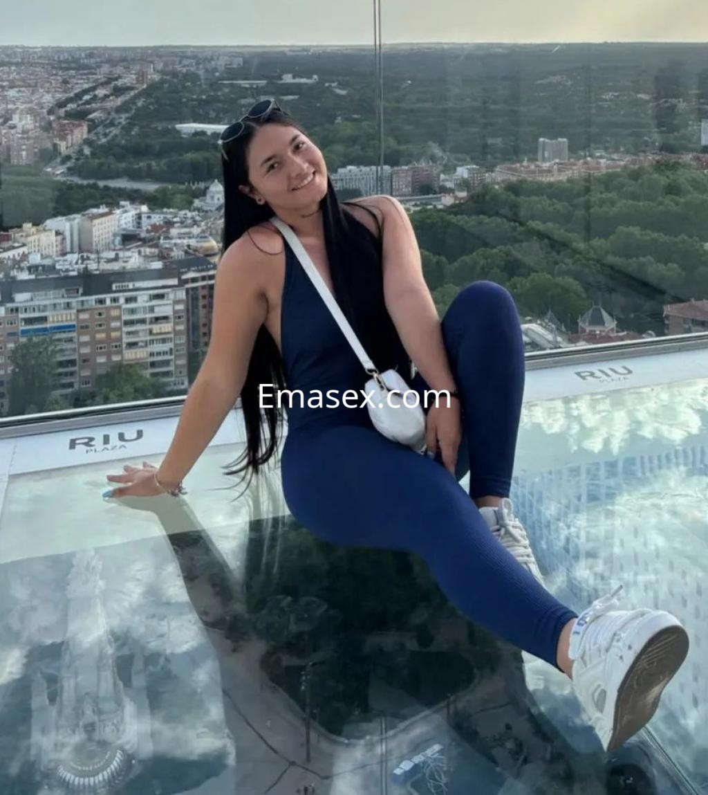 Escort en Zaragoza posando en un mirador con vista panorámica, vistiendo un mono azul y zapatillas blancas.