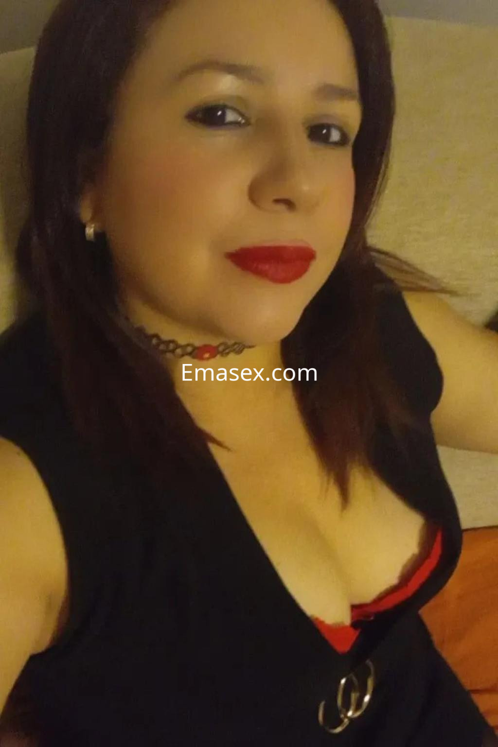 Escort con cabello castaño, luciendo un vestido negro con detalles rojos, posando en un entorno acogedor en Albacete.