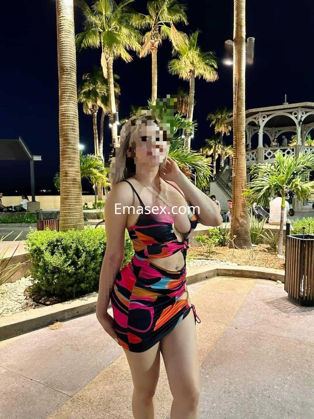 Escort en Albacete, vistiendo un vestido colorido y ajustado, posando en un entorno al aire libre con palmeras.