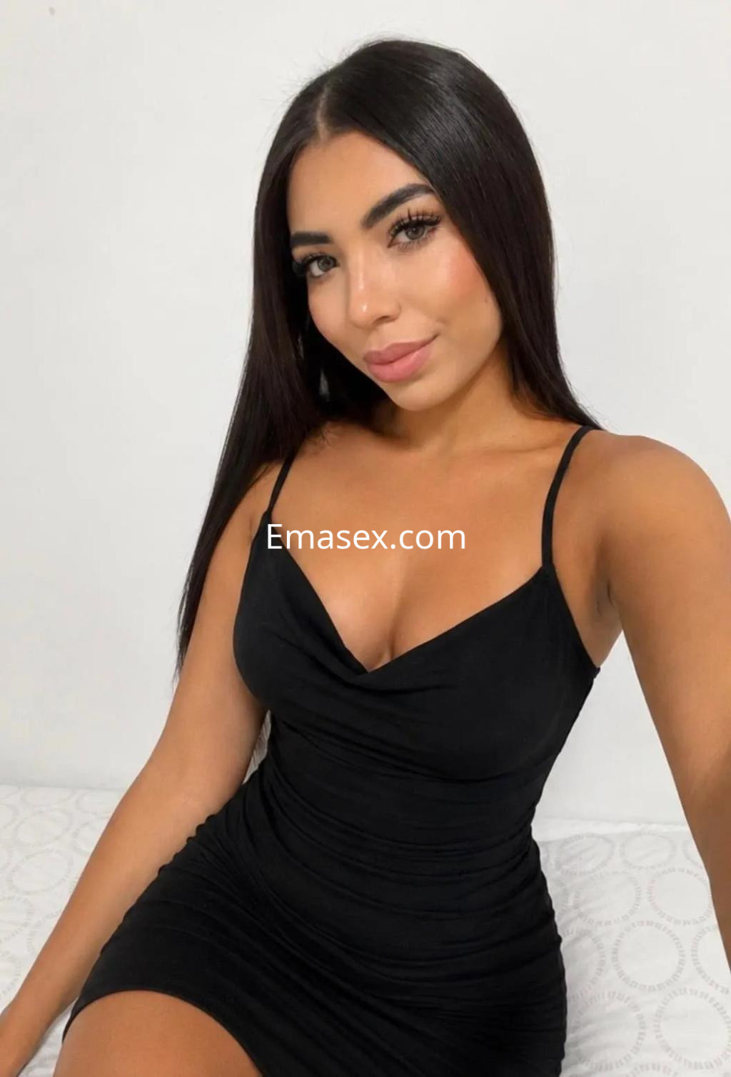 Escort en Alicante con vestido negro ajustado y tirantes, posando sobre una cama con fondo claro.