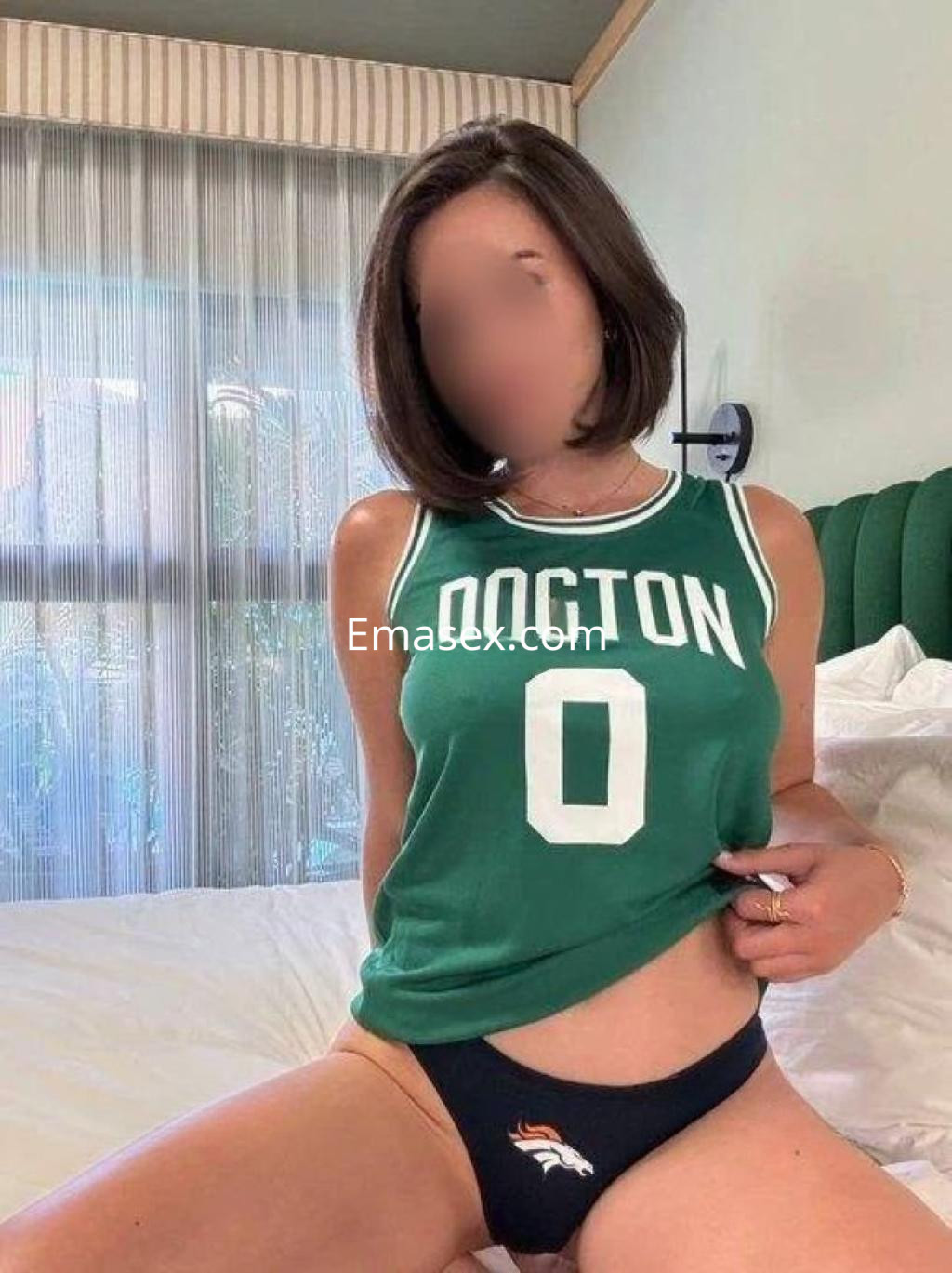 La puta en Ibiza viste una camiseta verde con el número 0 y una braguita negra, sentada en una cama.