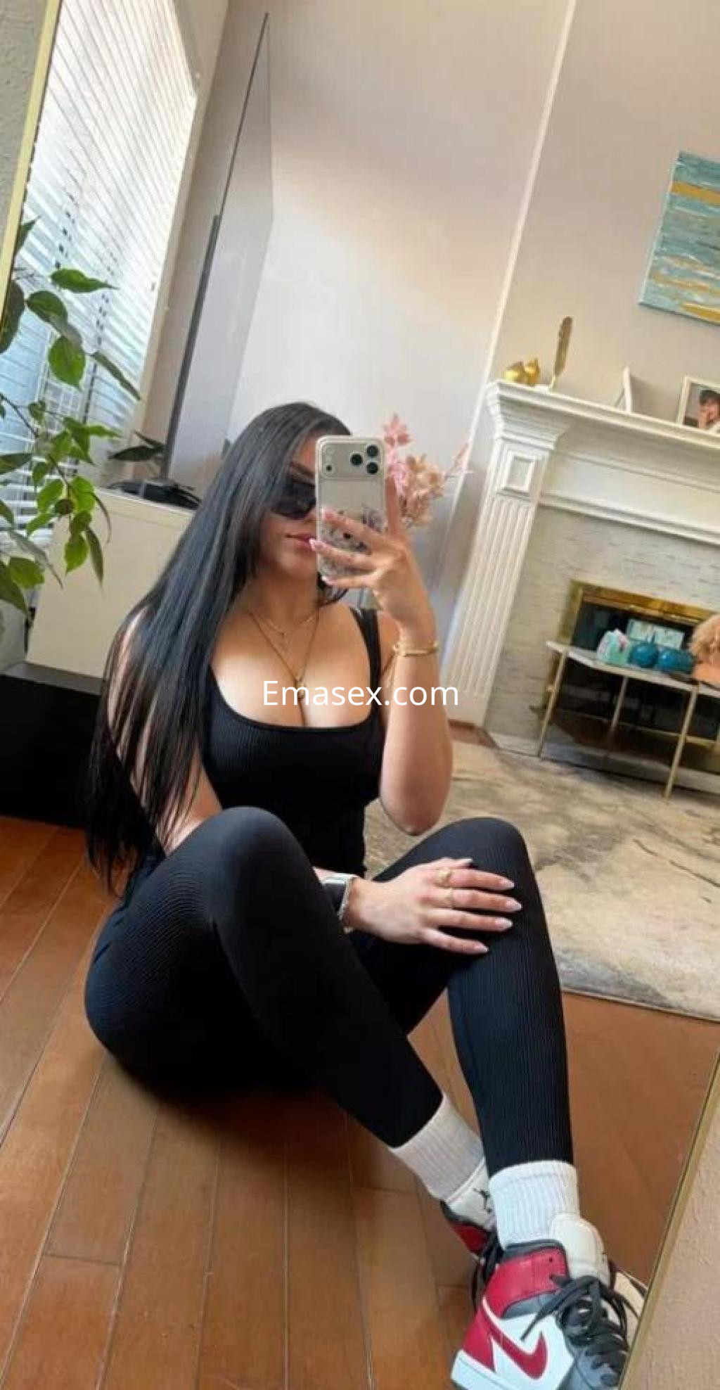 Escort en Lugo sentada en el suelo con ropa deportiva negra, gafas de sol y zapatillas blancas, mostrando su reflejo en el espejo.