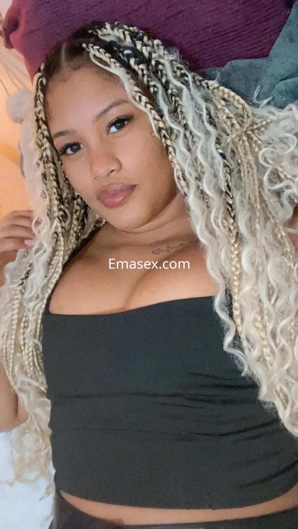 Escort en Vigo, con cabello trenzado de tonos rubios y oscuros, vistiendo una blusa sin mangas y recostada en un ambiente acogedor.