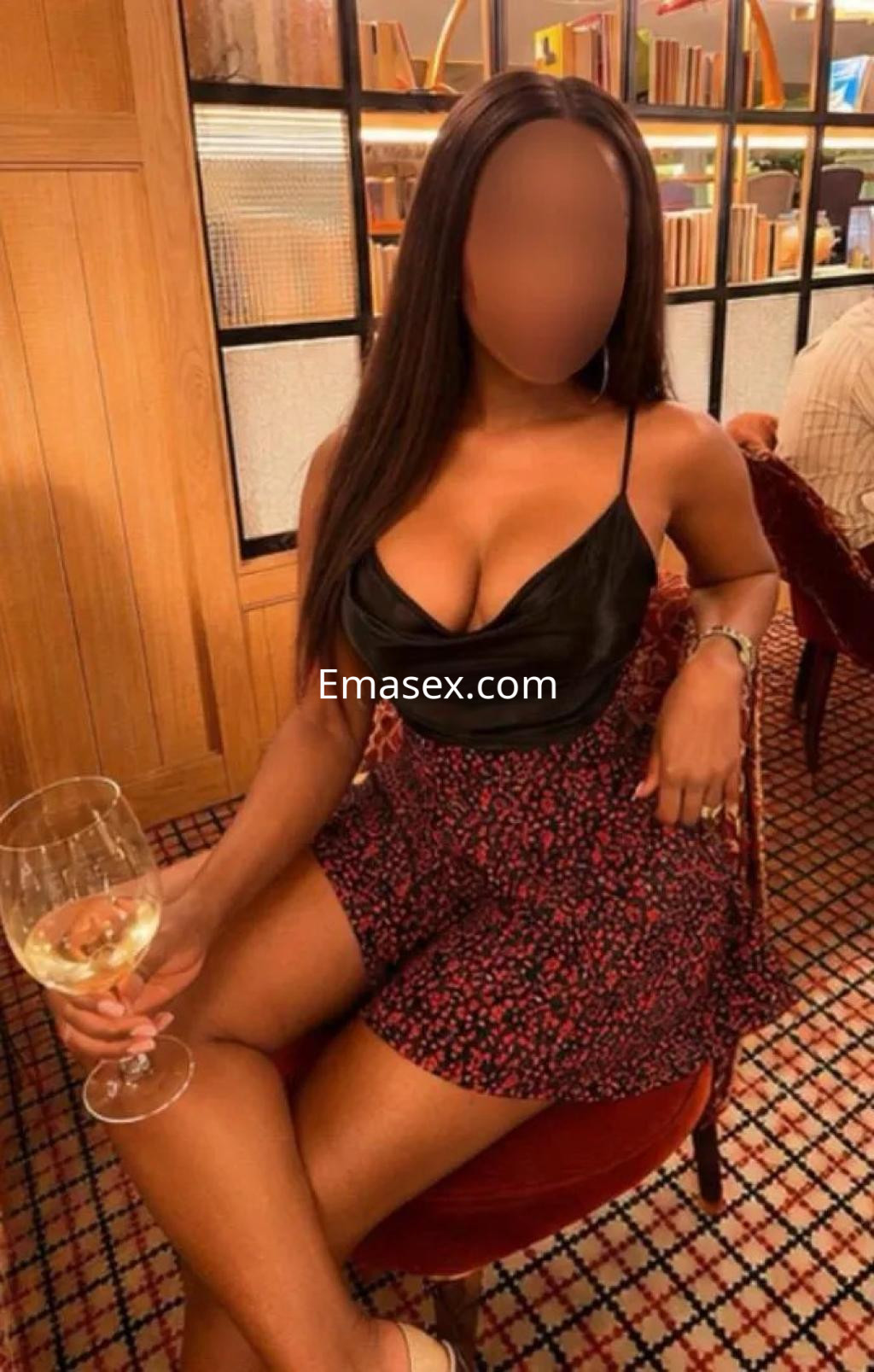 La puta en Vigo luce una blusa negra sin mangas y una falda corta con estampado, mientras sostiene una copa de vino.
