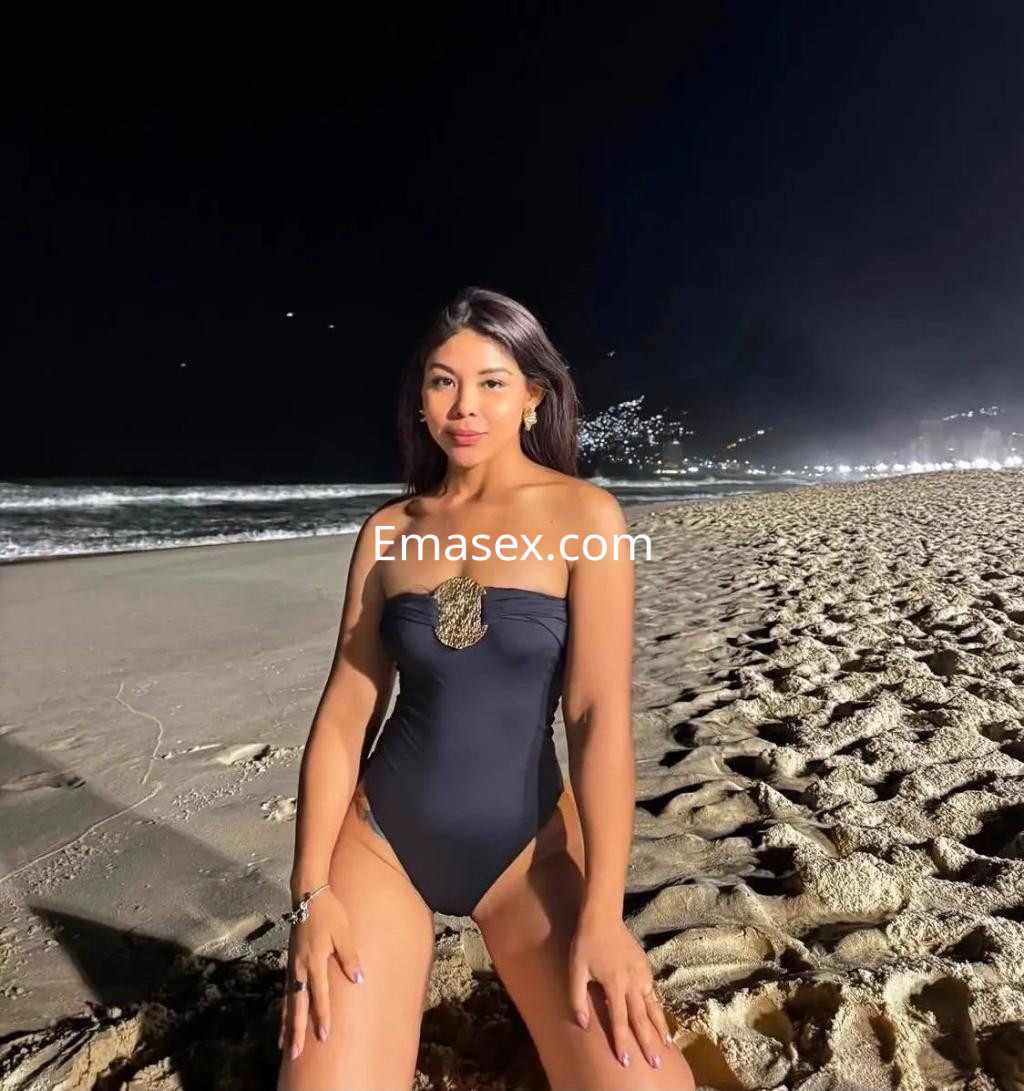 Escort posando en la playa de Valencia, con un bañador negro y el cabello largo suelto, bajo la luz nocturna.