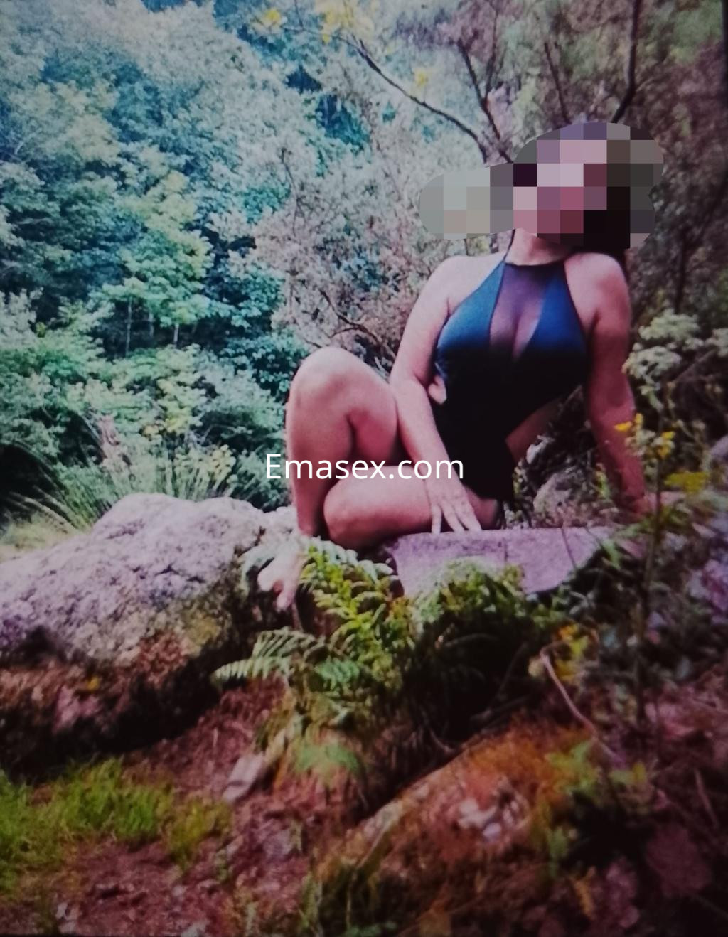 Puta en Donosti posando en un entorno natural, con un traje de baño negro y una pose sugestiva sobre una roca.