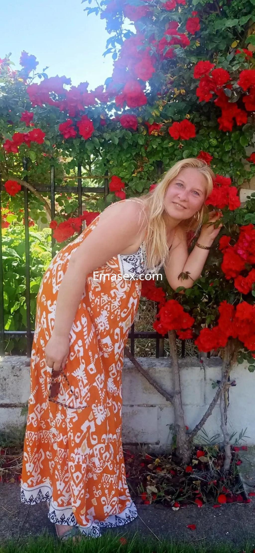 La puta posa sonriente junto a un arbusto de flores rojas, vistiendo un vestido largo y colorido en Oviedo.