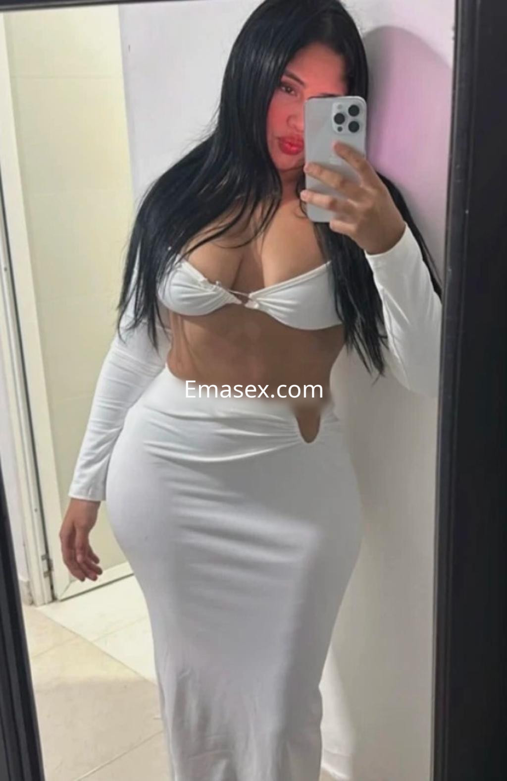 Puta en Gijón posando frente al espejo con un vestido blanco ajustado que destaca su figura y cabello largo.