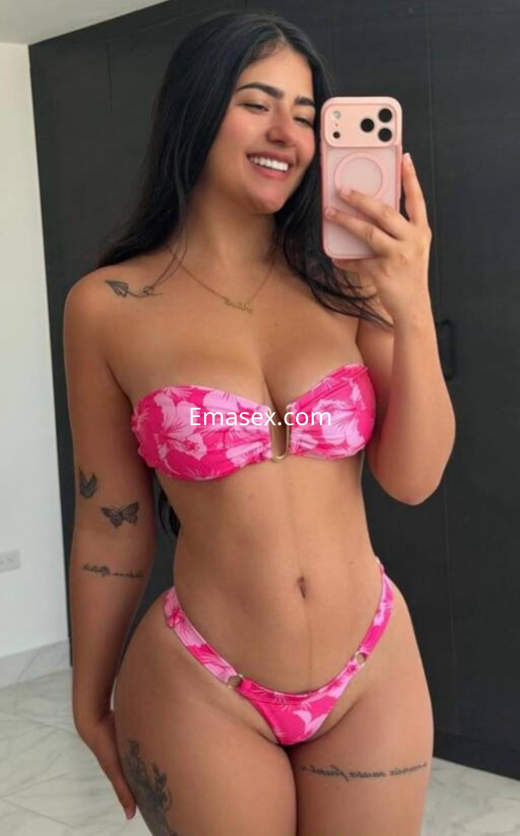 Escort en Pamplona posando con un bikini rosa, sonriendo y mostrando sus tatuajes en un fondo claro.